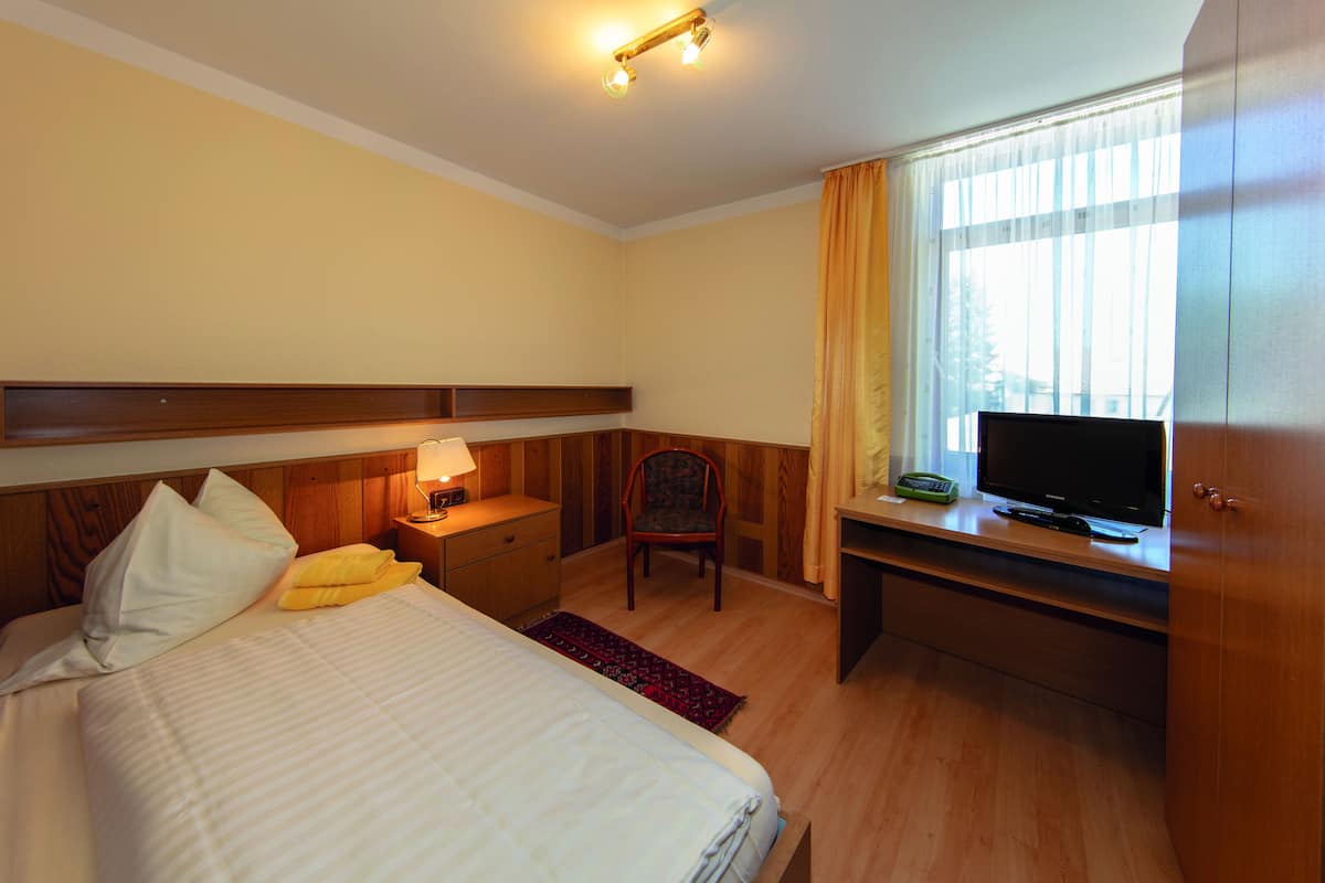Hotellihuone, jossa on sänky, työpöytä, jossa on televisio, tuoli, yöpöytä, jossa on lamppu, ja ikkuna, jossa on verhot.