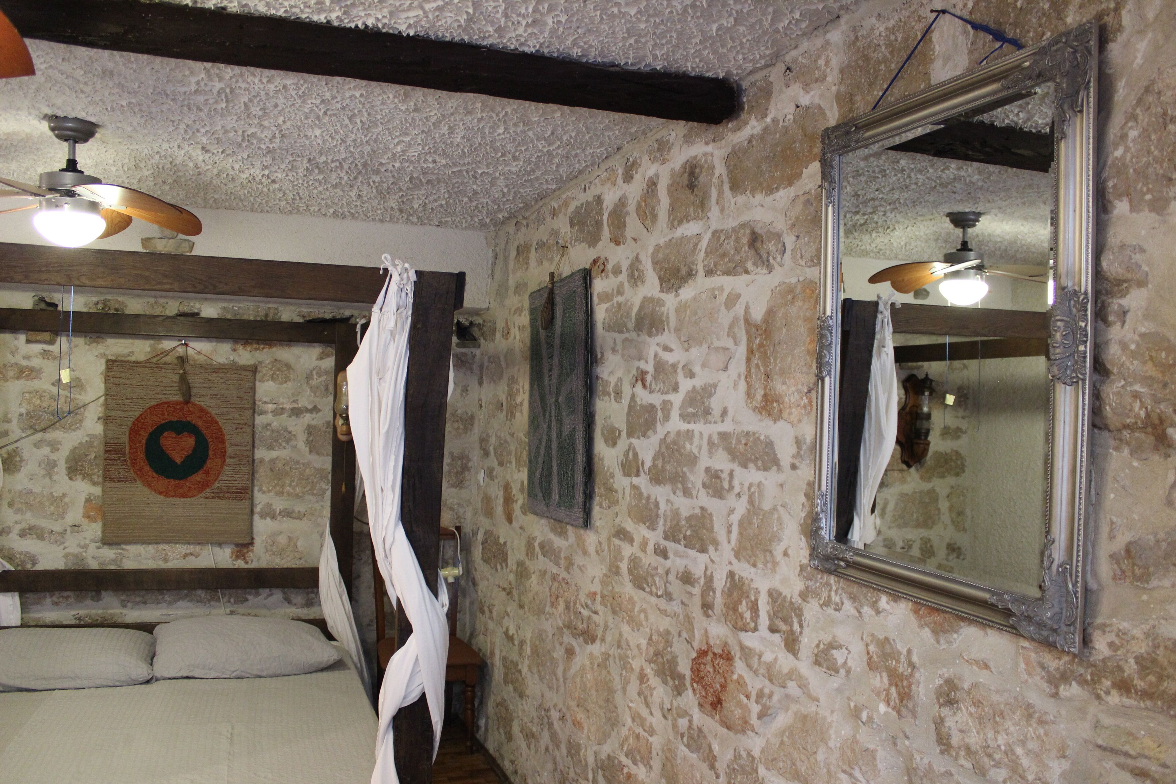 A room with stone walls, a bed with white bedding, a ceiling fan, and a large mirror.