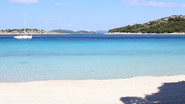 A beach with clear blue water, a sailboat, and a hilly landscape in the background. - Murter Voltera (Tisno)