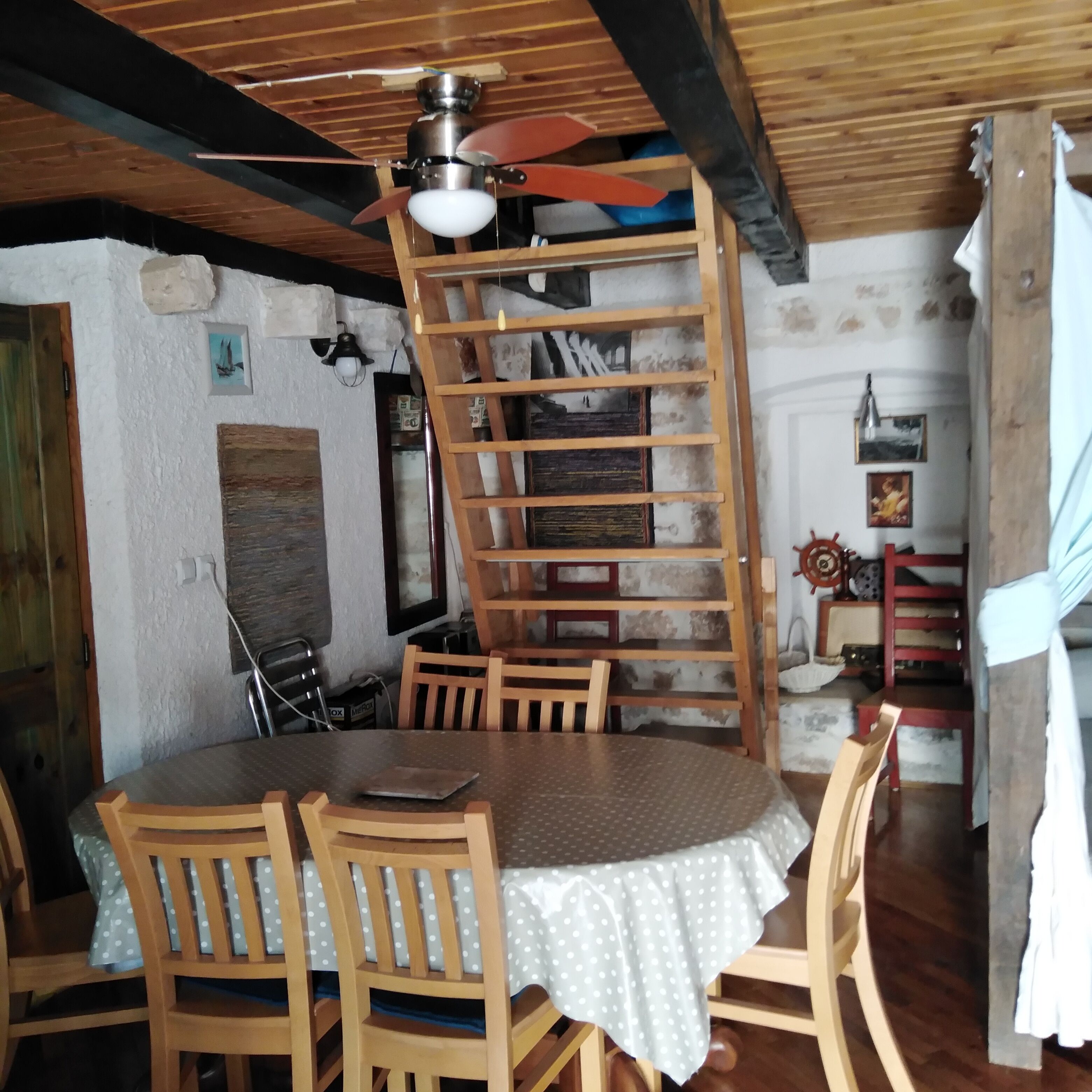 Un comedor con una mesa redonda, sillas de madera, un ventilador de techo y una estantería de escalera de madera.