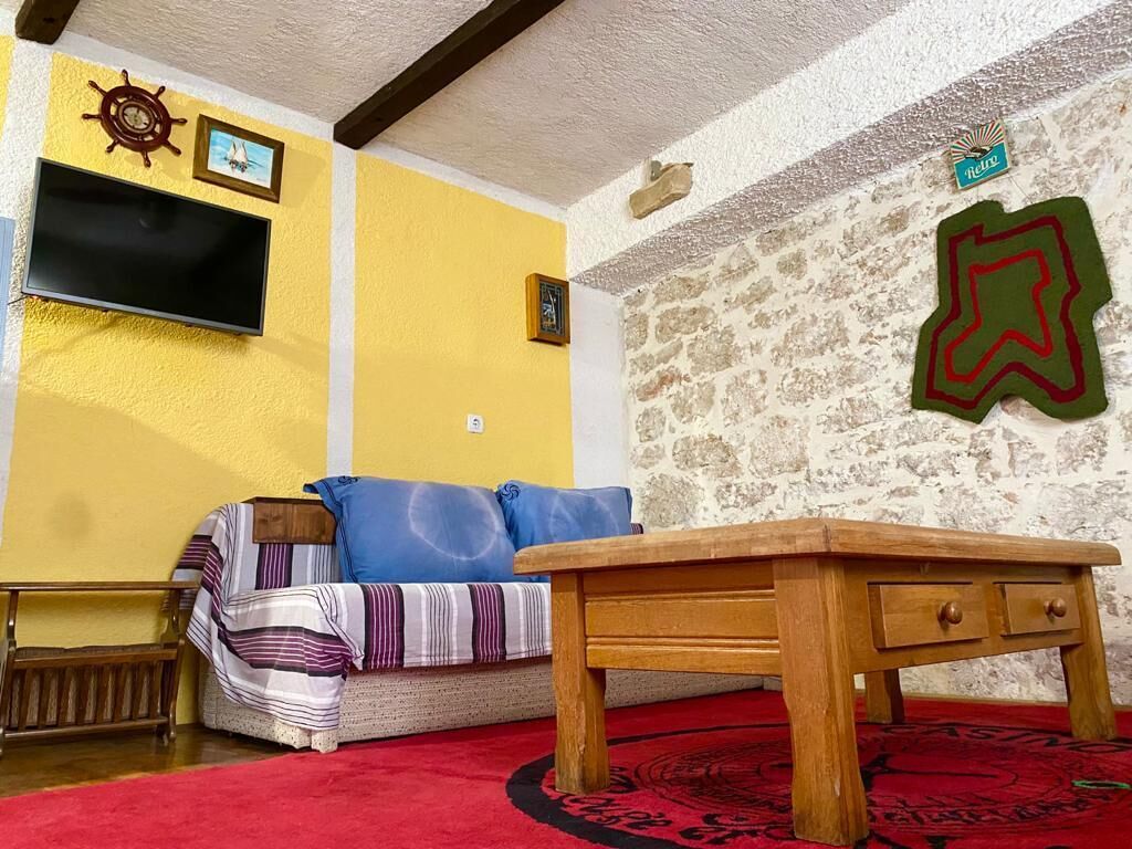 A cozy living room with a wooden coffee table, a sofa with striped cushions, a flat-screen TV mounted on a yellow wall, and a decorative wall hanging.