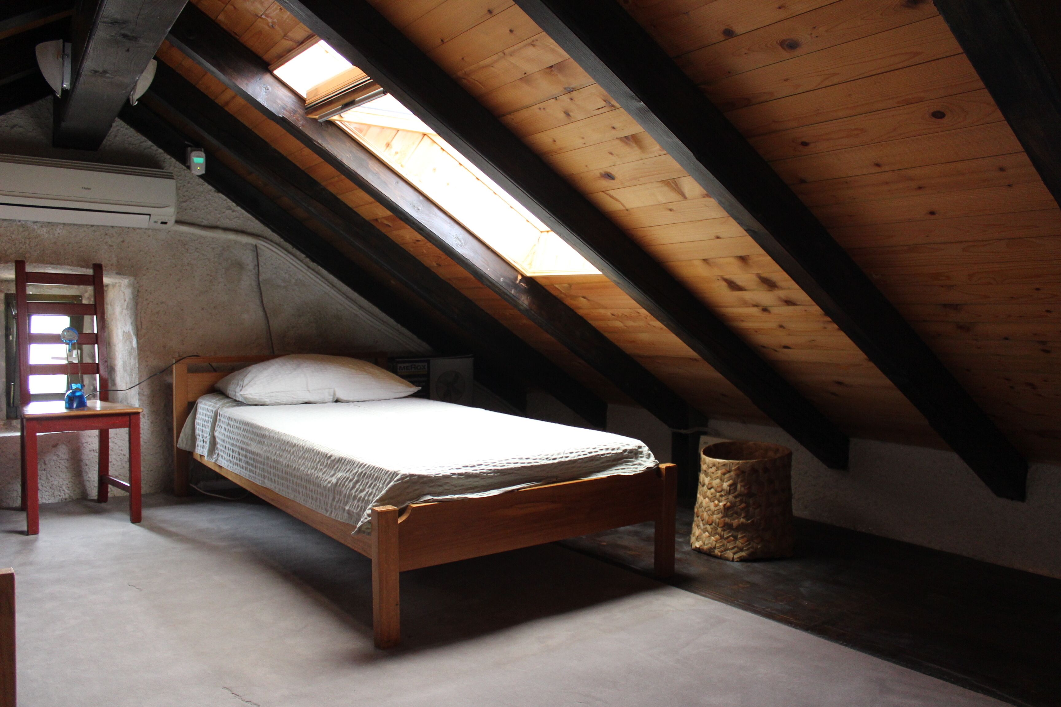 Una cama individual en una habitación con techo inclinado y tragaluz, una silla y un recipiente de piedra.