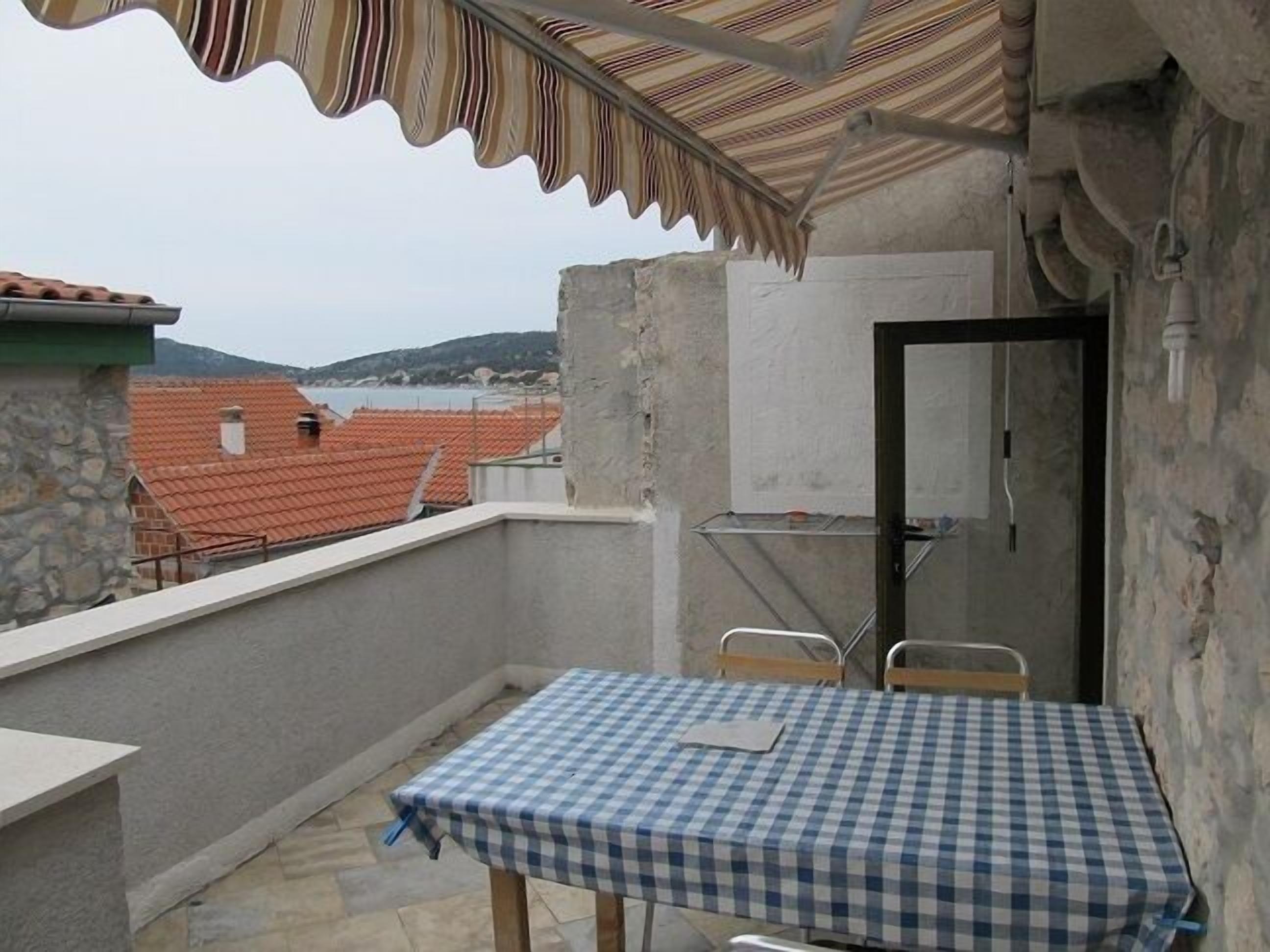 A balcony with a checkered table and chairs, a stone wall, and a view of red-roofed buildings and a body of water in the background.