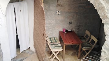 Una habitación rústica con pared de piedra, una mesa de madera con sillas, un estante con objetos y una cortina blanca.