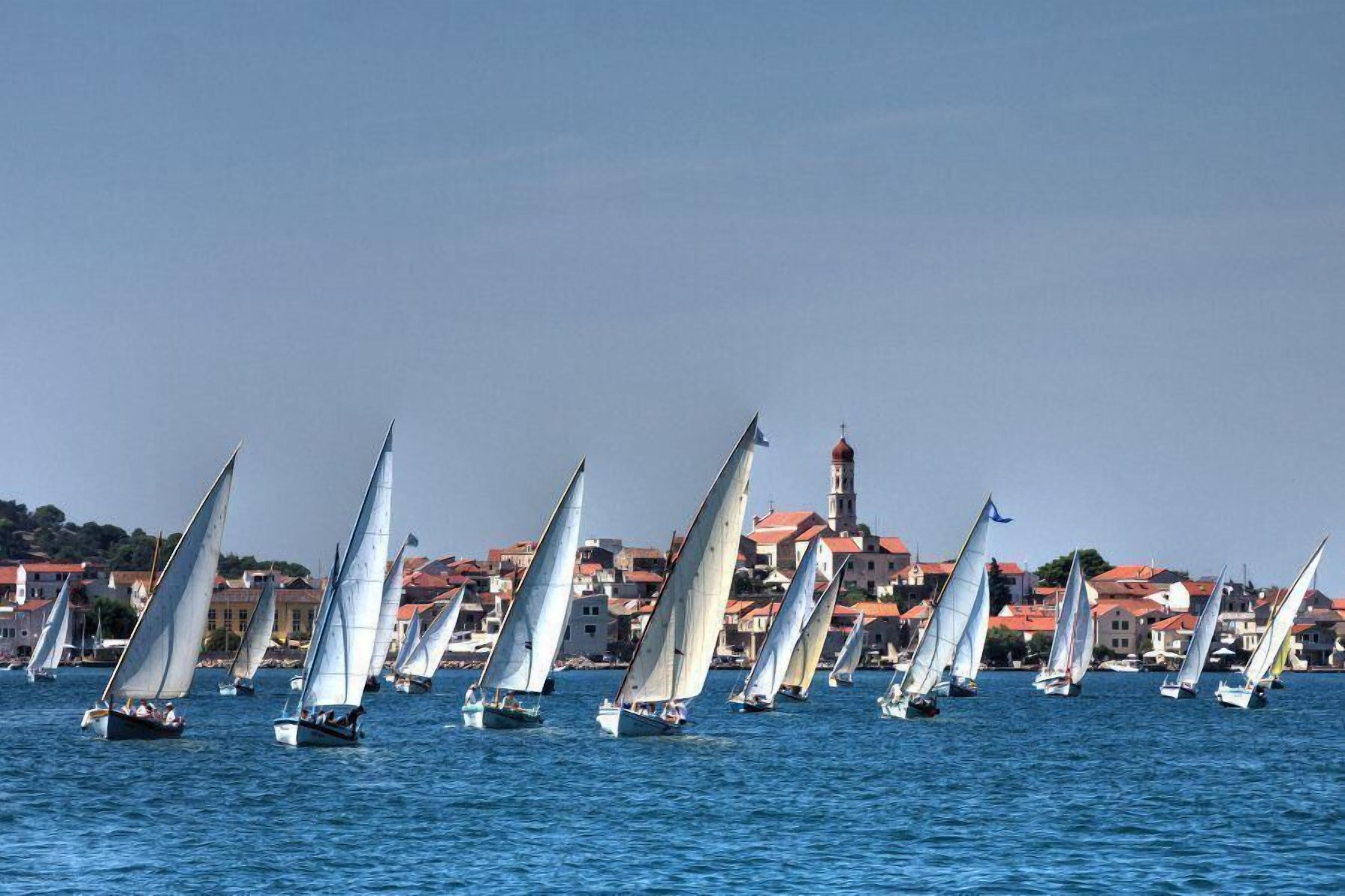 Eine Küstenstadt mit Segelbooten auf dem Wasser, Häusern mit roten Dächern und einem Turm im Hintergrund.