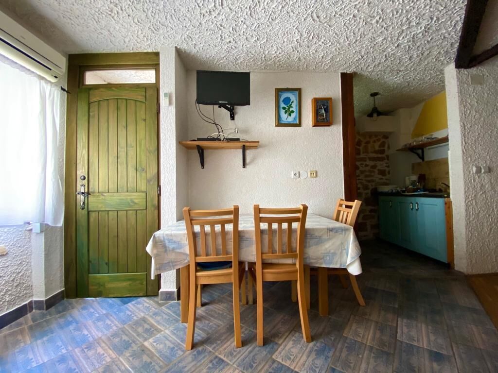 Un comedor con una mesa y sillas de madera, una puerta verde, un televisor en la pared y una cocina al fondo.