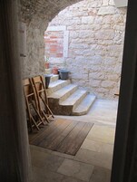 Un patio con paredes de piedra, una escalera de madera apoyada en una pared, escalones de piedra y macetas con plantas.