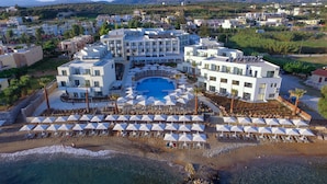 An aerial view of a resort with a central pool, multiple buildings, and a beach area with umbrellas and sun loungers. - Harmony Rethymno Beach Hotel (Rethymno)
