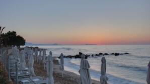 A beach with rows of sunbeds and umbrellas, a calm sea, and a sunset in the background. - Harmony Rethymno Beach Hotel (Rethymno)