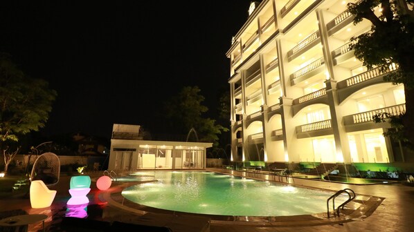 Vista nocturna de un hotel con piscina, mobiliario exterior y un edificio de varios pisos con balcones.