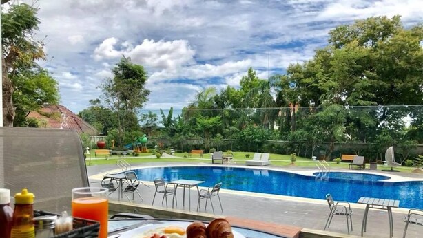 A breakfast table with a plate of food, a cup of coffee, and a bottle of juice, overlooking a swimming pool with lounge chairs and greenery. - The Park Vientiane (Vientiane)