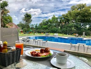 Una mesa de desayuno con un plato de comida, una taza de café y una botella de jugo, con vistas a una piscina con sillas de descanso y follaje.