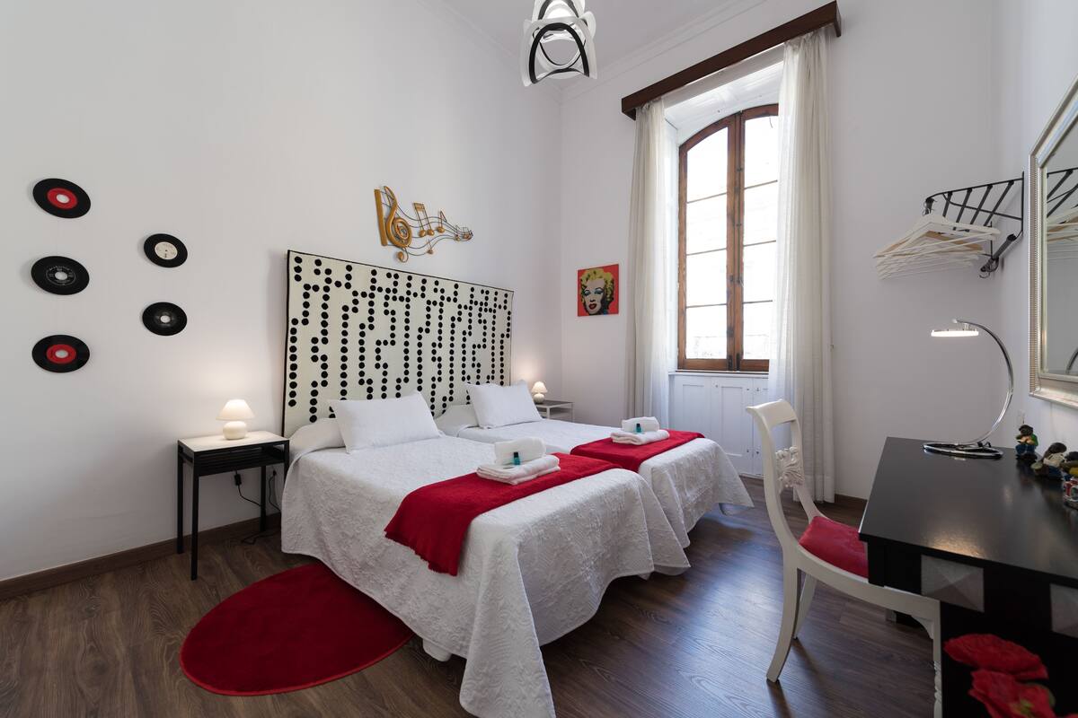 A hotel room with two beds, a red rug, a desk, a mirror, and a window with curtains.