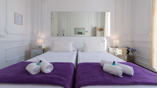 A hotel room with two beds, each with a purple blanket and rolled towels. There are bedside tables with lamps and a mirror above the beds. - Casa Cinzia (Las Palmas de Gran Canaria)