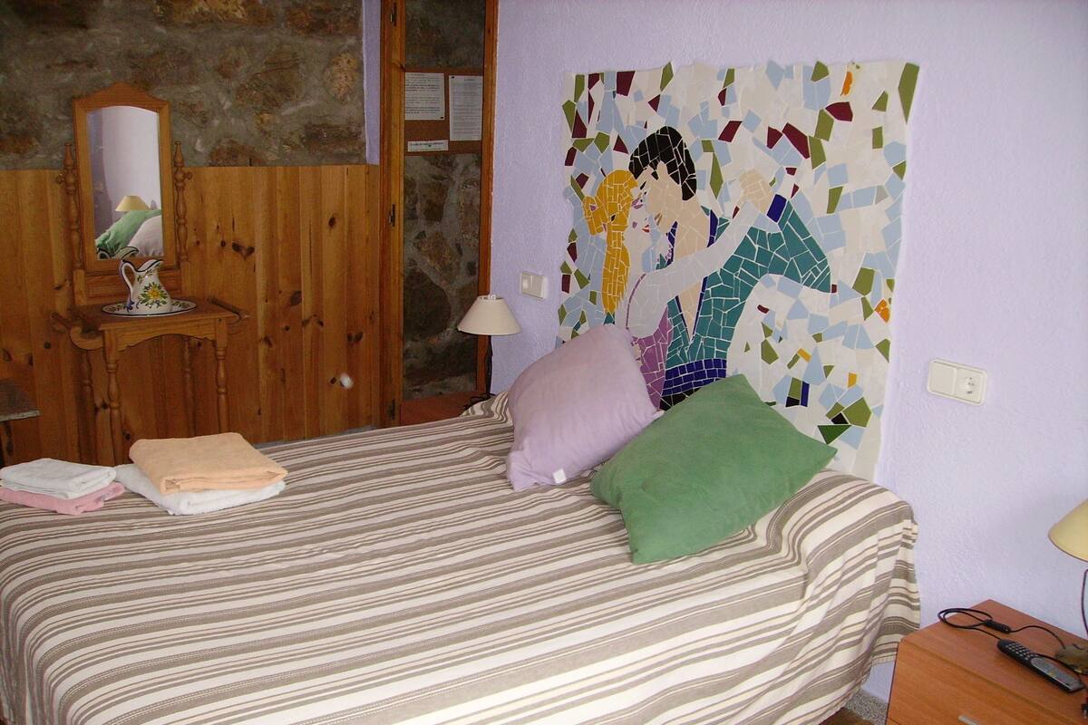 A bed with striped bedding, a green pillow, and a pink pillow. There is a wooden nightstand with a lamp and a remote control. A mosaic artwork is on the wall above the bed.