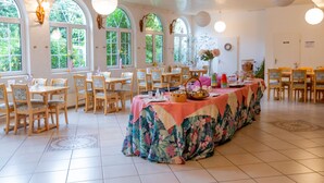 Uma sala de jantar espaçosa com várias mesas e cadeiras, grandes janelas com arcos e arandelas decorativas.