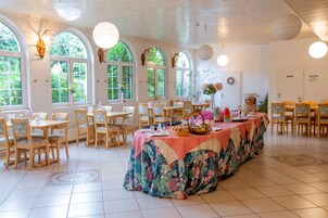 A spacious dining area with multiple tables and chairs, large windows with arched tops, and decorative wall sconces.