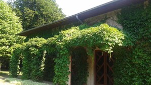 A building with a covered entrance and a wooden door, surrounded by lush greenery and a gravel path. - La Passagère (Hauterive)