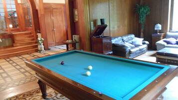 Une table de billard dans une pièce dotée d’un tapis à motifs, de meubles en bois et d’une vue sur l’extérieur, visible à travers une porte en arc de cercle.