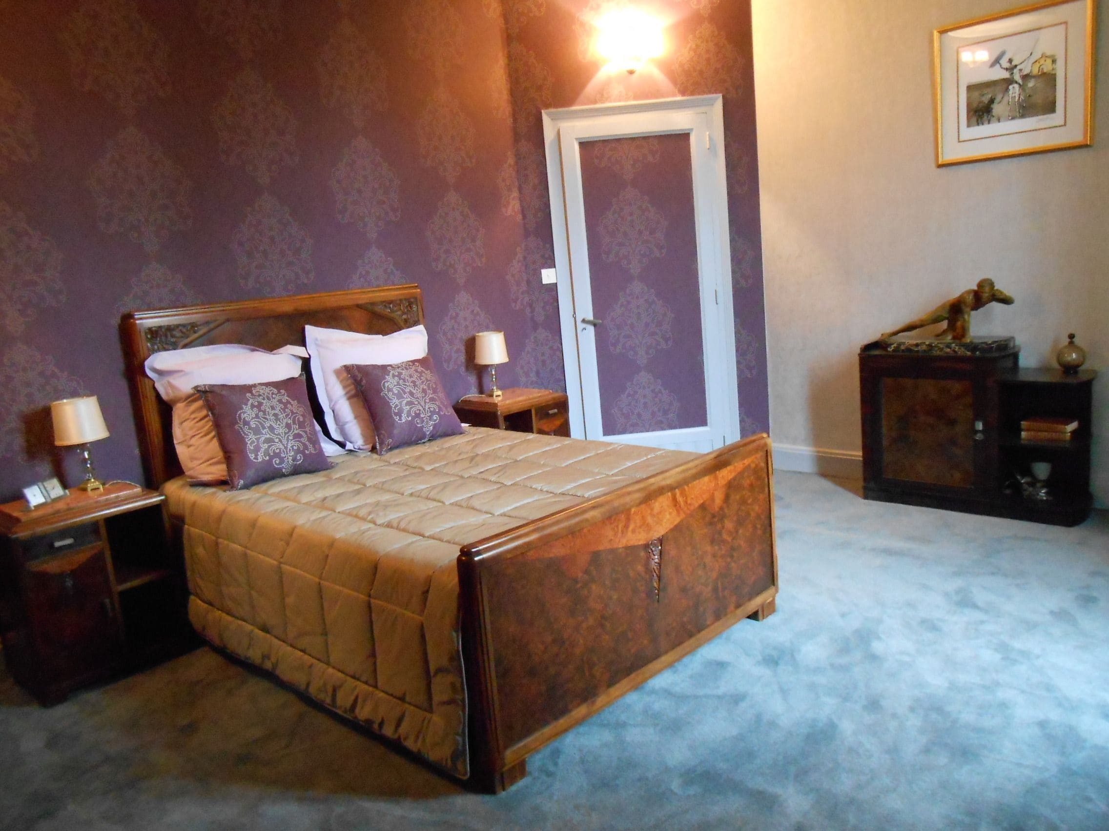 A neatly made bed with a wooden headboard, two bedside tables with lamps, a door, and a small cabinet with a statue on top.
