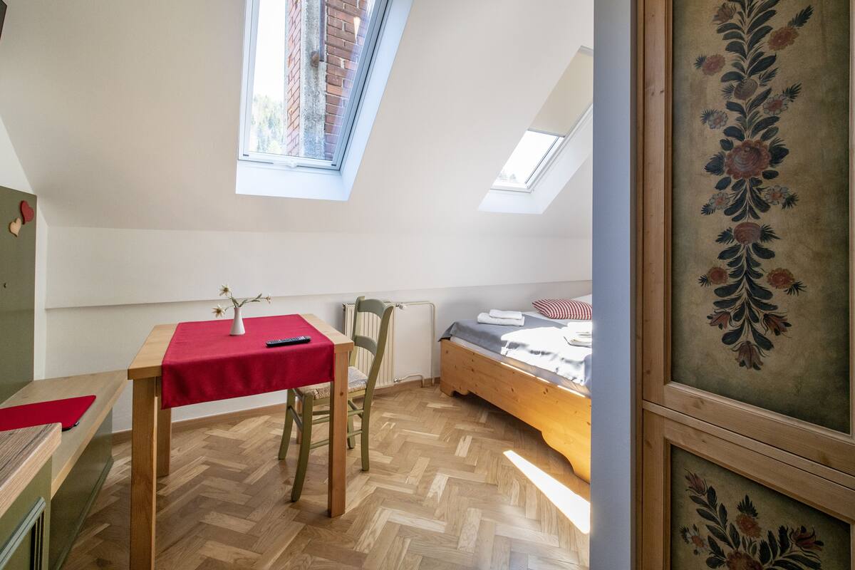 Una habitación con cama elevada, una mesa pequeña con mantel rojo, una silla y un radiador. Se ve una ventana con vista a un muro de ladrillo y un árbol.