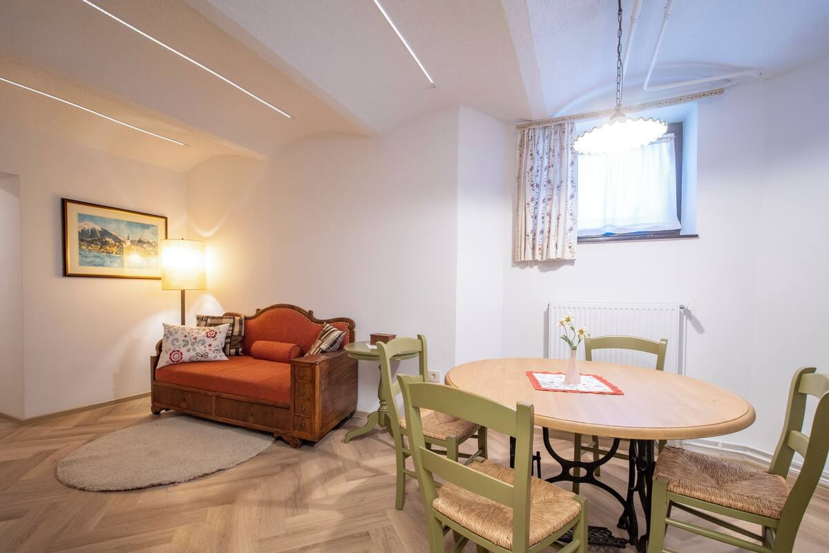Una habitación acogedora con piso de madera en diseño espiga, un sofá de madera con un cojín rojo, una mesa redonda de madera con cuatro sillas verdes, y una lámpara de mesa.