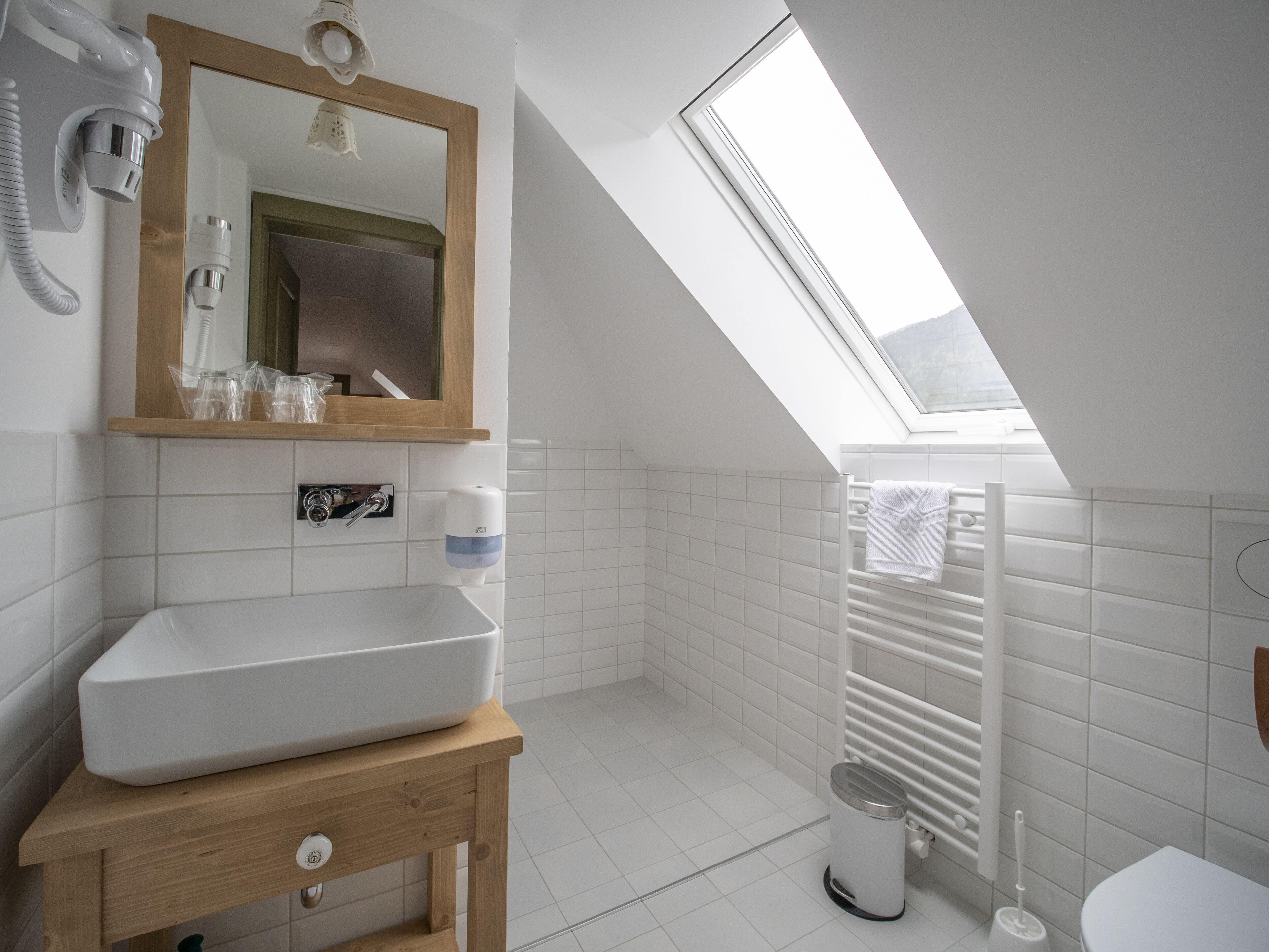 Un baño moderno con tragaluz, baldosas blancas, tocador de madera, inodoro y barra para toallas.