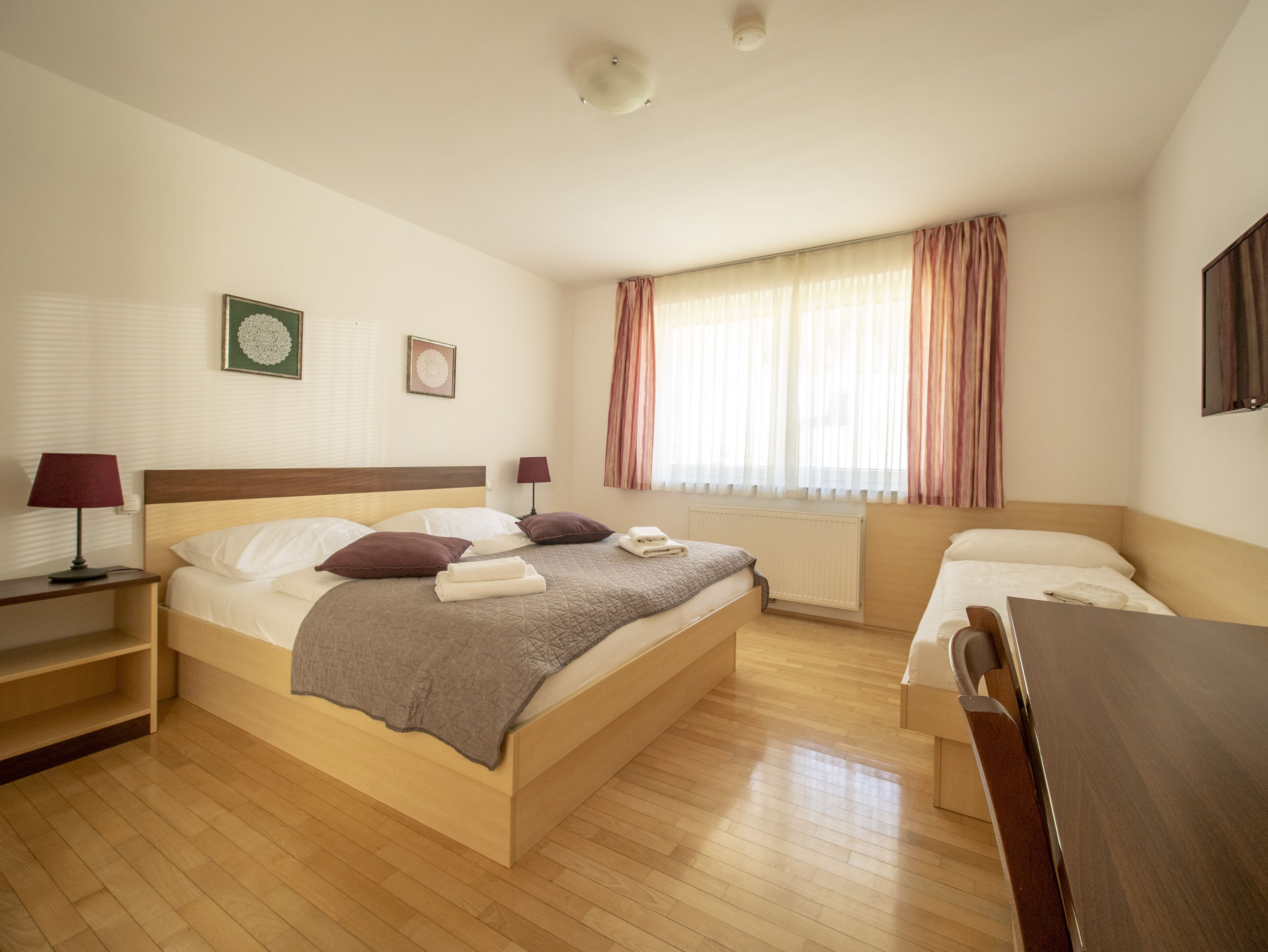 A hotel room with a large bed, a bedside table, a chair, a window with curtains, and a radiator.