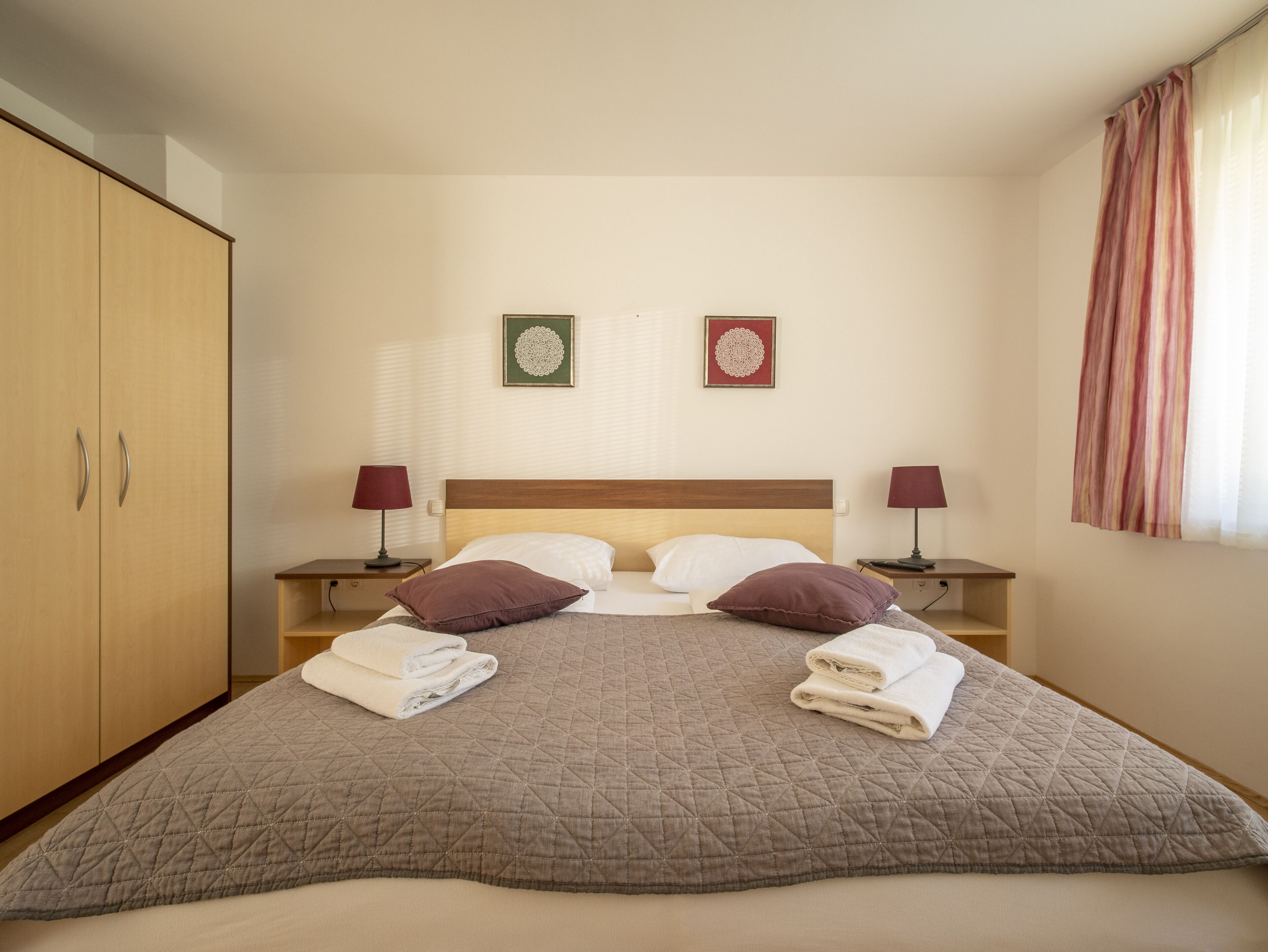 Een net opgemaakt bed met een gewatteerde sprei, twee kussens en opgevouwen handdoeken. Twee nachtkastjes met lampen, een garderobe en twee ingelijste foto's aan de muur.