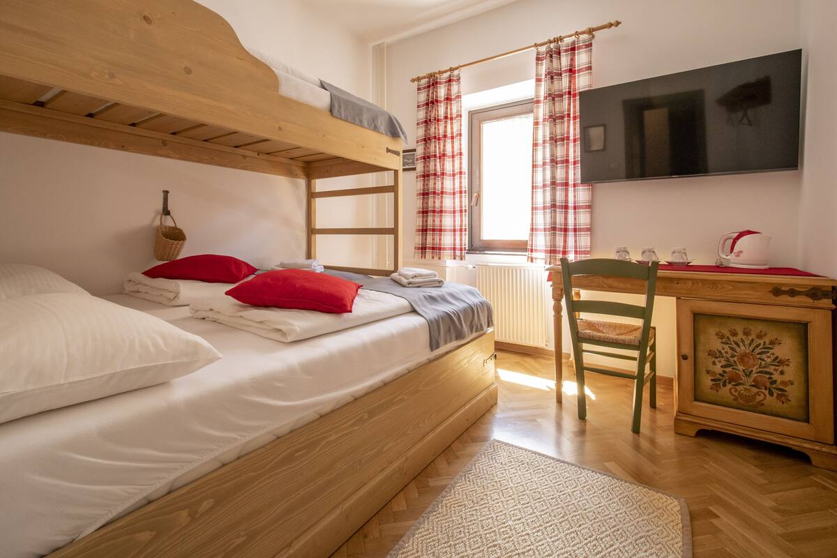 Una cama litera con ropa de cama blanca y almohadas rojas, un escritorio de madera con una silla, un televisor en la pared y una ventana con cortinas a cuadros.