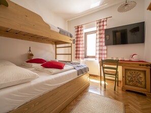 Una cama litera con ropa de cama blanca y almohadas rojas, un escritorio de madera con una silla, un televisor en la pared y una ventana con cortinas a cuadros.