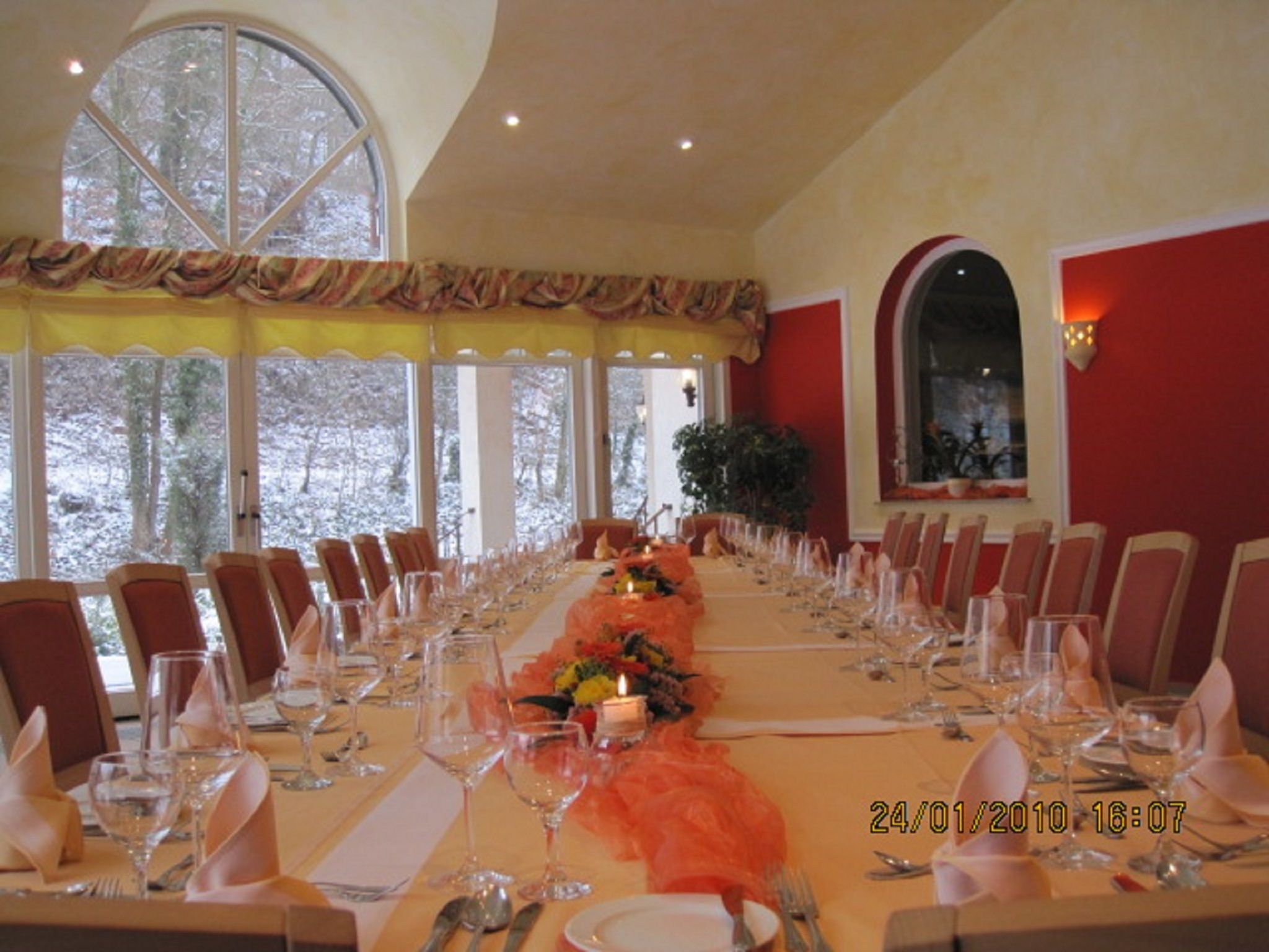 A long dining table set for a formal event with chairs on both sides, large windows with a view of trees, and a red accent wall.