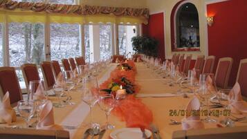 A long dining table set for a formal event with chairs on both sides, large windows with a view of trees, and a red accent wall.