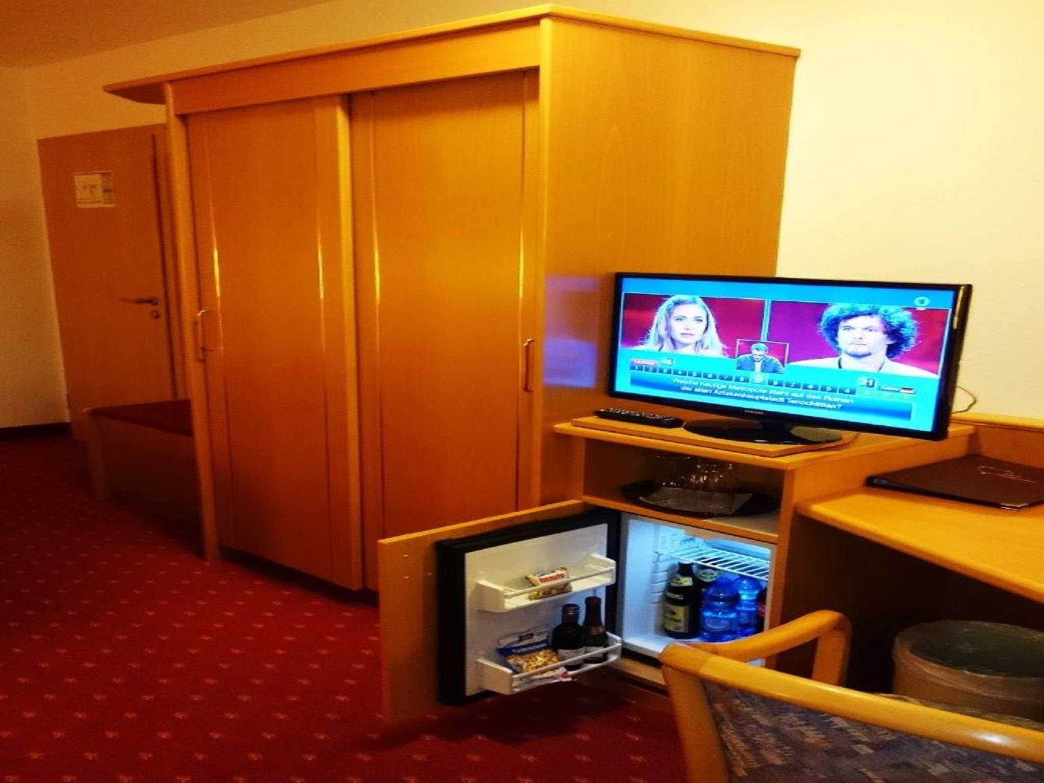 Habitación de hotel con televisión mostrando un noticiero, un mini refrigerador con bebidas, una computadora portátil y un armario de madera.