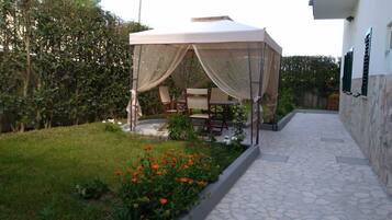 A covered outdoor seating area with a table and chairs, surrounded by a garden with orange flowers and a stone pathway.
