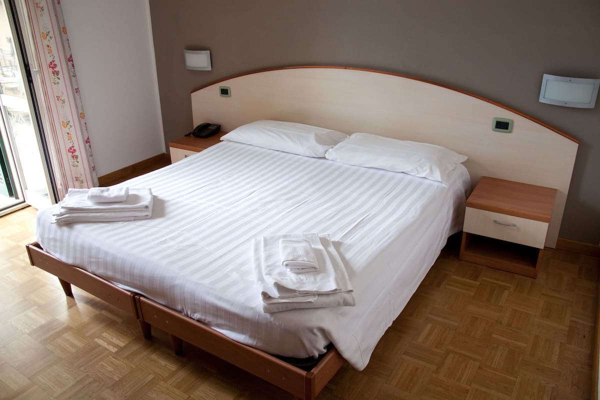 Cama bem arrumada com lençóis brancos e toalhas dobradas, cabeceira de madeira, mesa de cabeceira com abajur e criado-mudo com telefone.