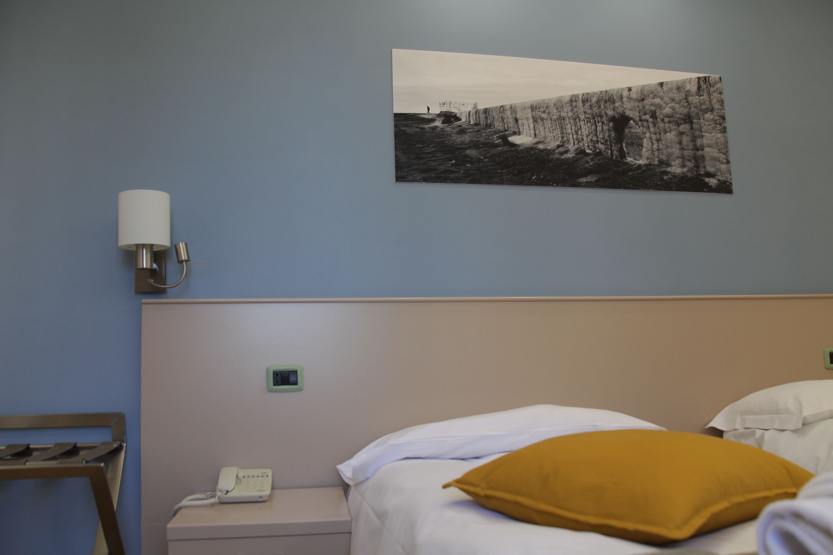 A hotel room with a bed, a nightstand with a phone, a wall-mounted lamp, and a picture of a coastal scene.