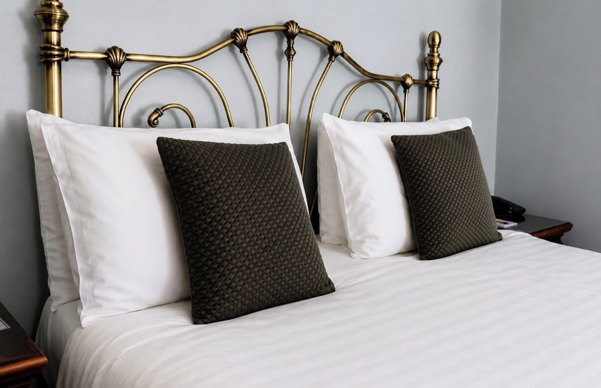 Una cama con cabecera metálica, almohadas blancas y verde oscuro, y una colcha blanca.