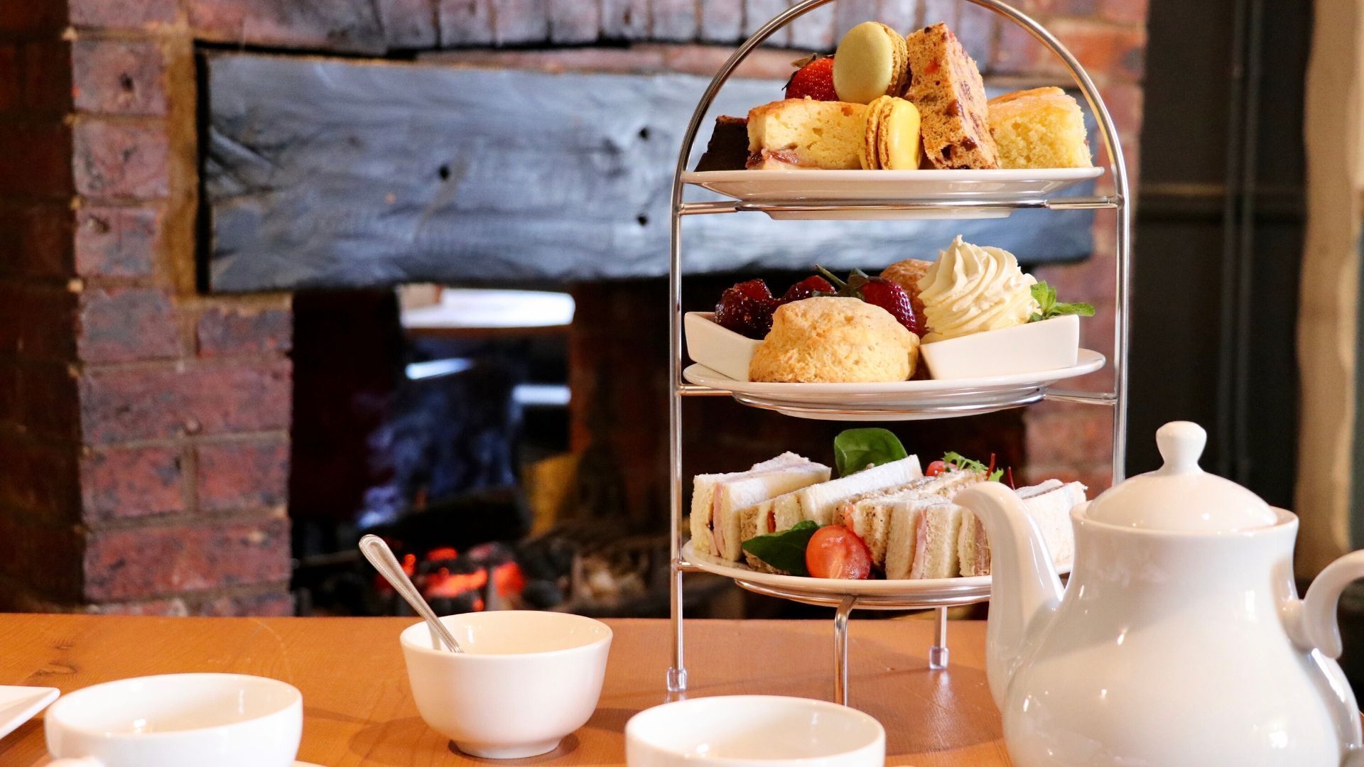 A tiered dessert stand with various pastries and sandwiches, a teapot, and cups on a wooden table in front of a brick wall.