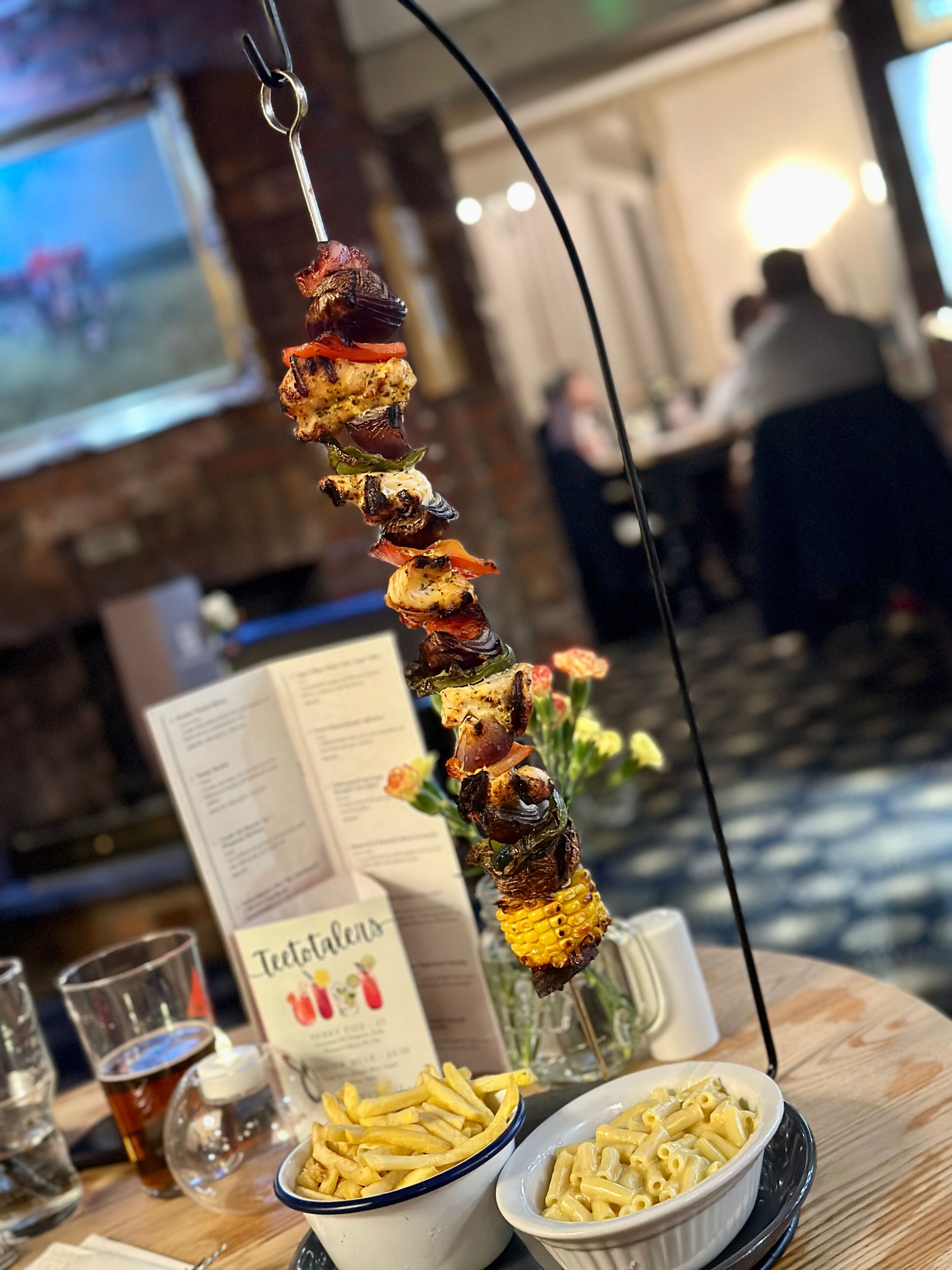 A skewer of grilled vegetables and meat, a menu, a bowl of fries, and a bowl of macaroni and cheese on a table in a restaurant.