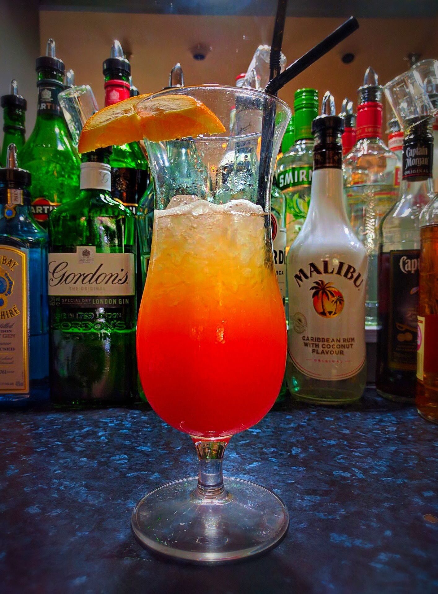 A cocktail glass with a gradient color, a slice of orange, and a straw, placed on a bar counter surrounded by various liquor bottles.
