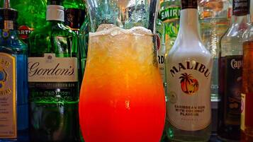 A cocktail glass with a gradient color, a slice of orange, and a straw, placed on a bar counter surrounded by various liquor bottles.