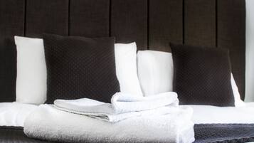 Una cama bien hecha con ropa de cama blanca y gris oscuro, una toalla blanca doblada y un almohadón texturizado gris oscuro.