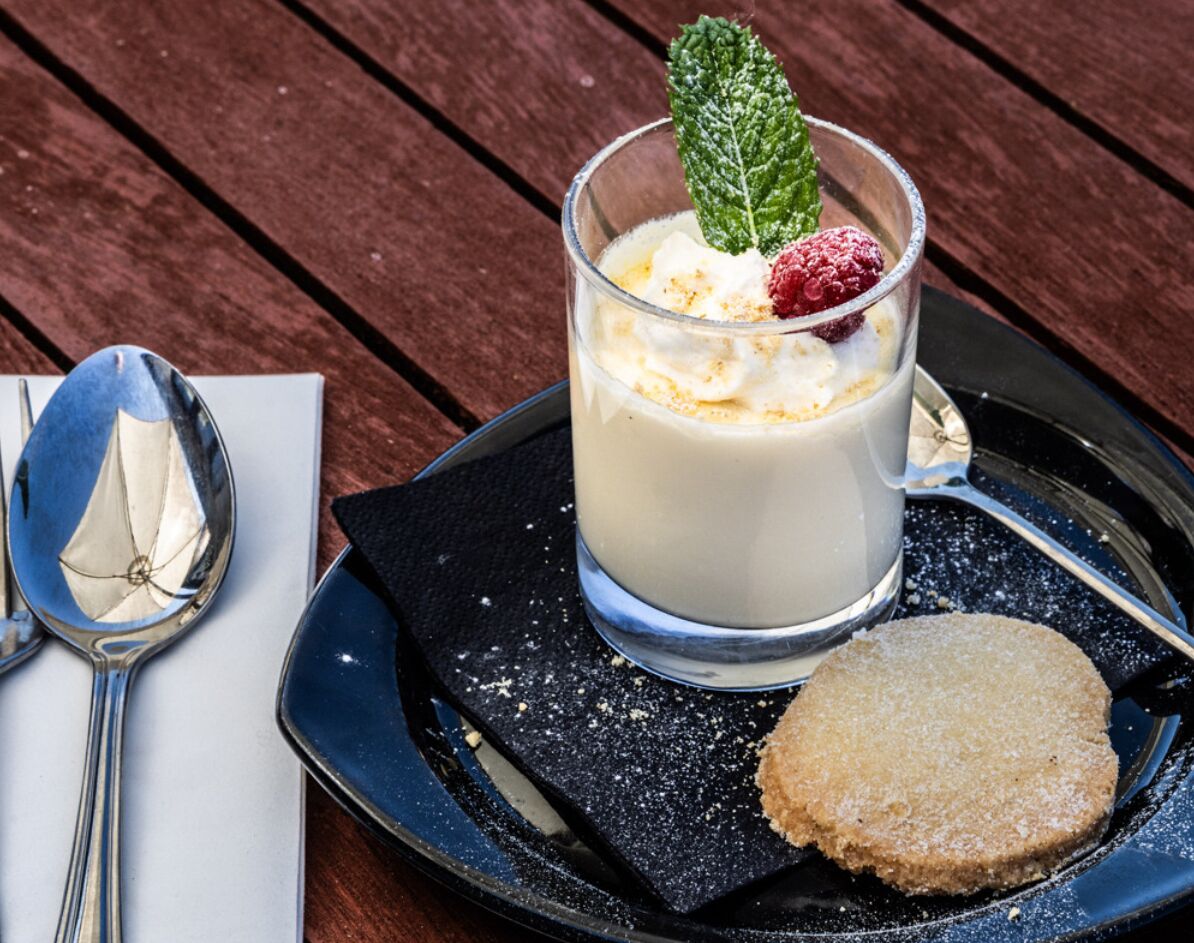 Un postre que incluye un vaso de bebida cremosa, crema batida, una frambuesa y una hoja de menta, acompañado de una galleta sobre un plato oscuro.
