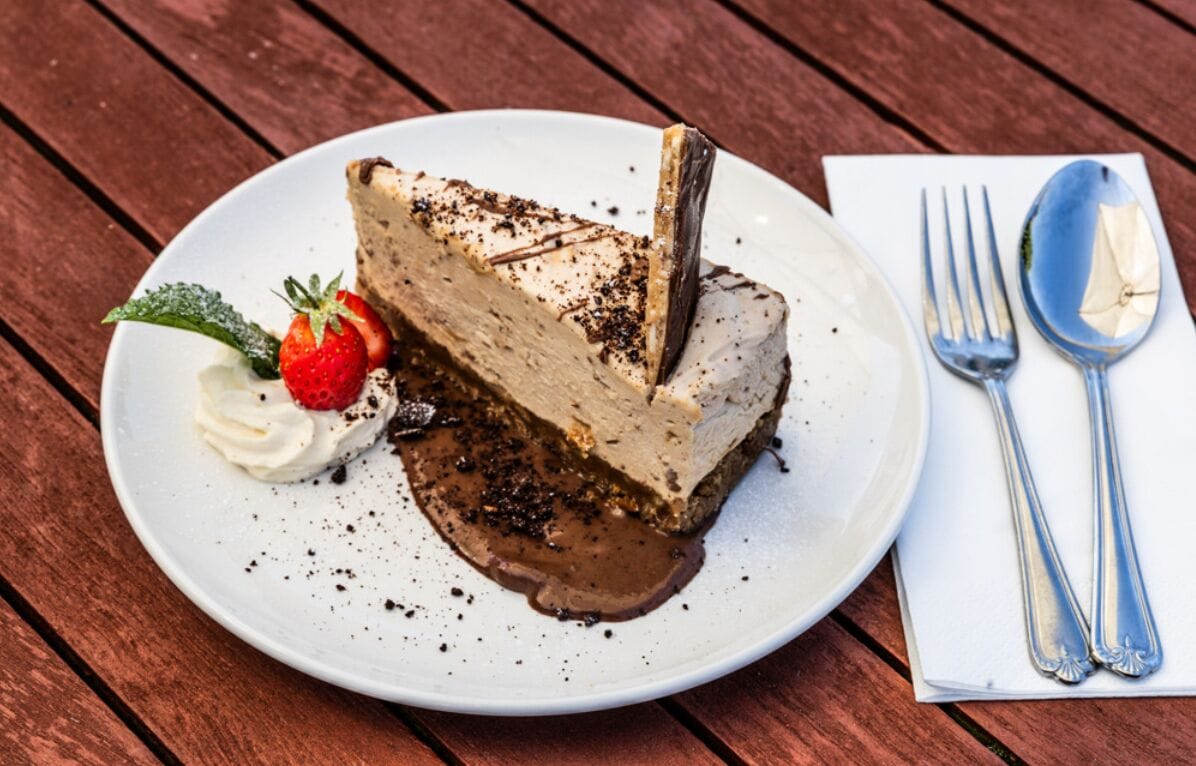 A slice of cheesecake on a white plate with a strawberry and mint garnish, accompanied by a fork and spoon on a napkin.