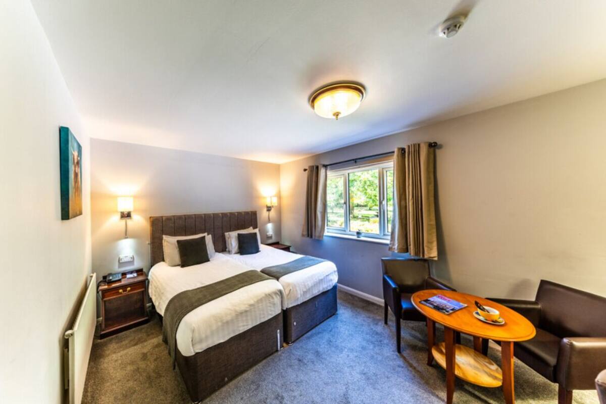 Ein Hotelzimmer mit Doppelbett, zwei Nachttischen, einem kleinen Tisch mit Stuhl, einem Fenster mit Vorhängen und einer Deckenleuchte.