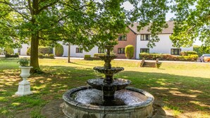 A circular stone fountain with water cascading down its tiers, surrounded by a well-maintained lawn and a large tree providing shade. - The Boars Head Hotel (Ashbourne)