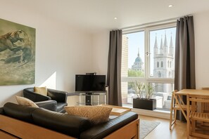 A modern living room with a leather sofa, a flat-screen TV, and a dining area with a wooden table and chairs. A large window offers a view of a historic building. - Still Life St. Paul's Executive (London)