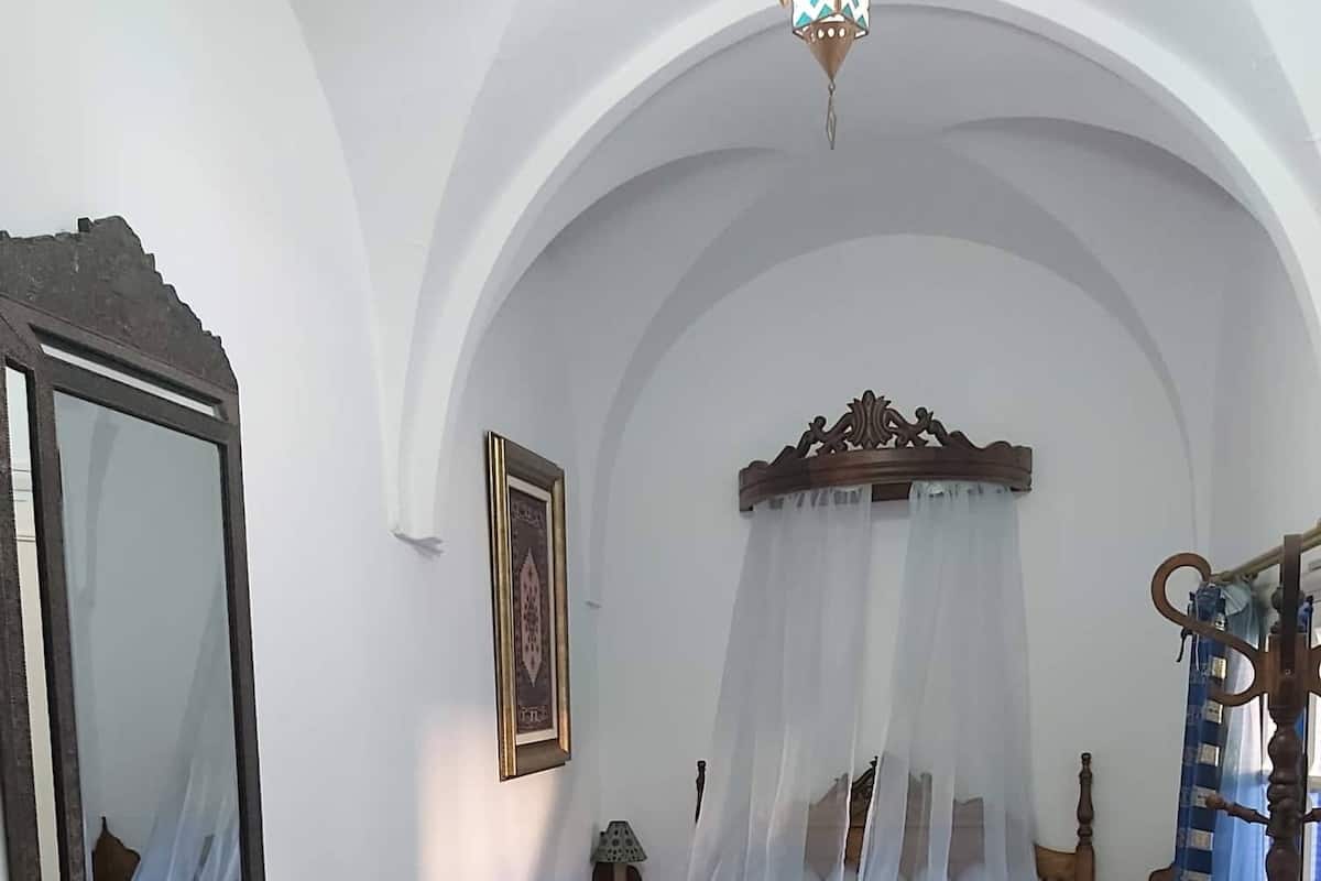 A bedroom with a bed under a canopy, a bedside table, a mirror, a framed picture, and a wooden chair.