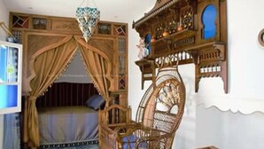 A room with a bed under a canopy, a wicker chair, and a shelf with decorative items. - Dar El Goulli (Sousse)